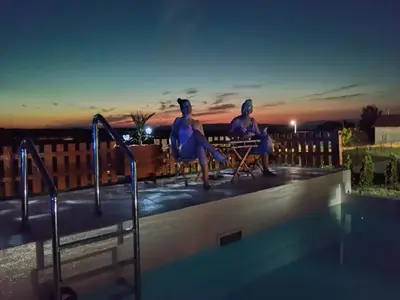 two women sitting next to a swimming pool at night at Vikendica sa bazenom Čarolija Kruševac in Kruševac