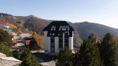a white building with a black roof with mountains in the background at Lawa Apartments Kopaonik in Cajetina