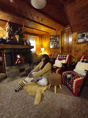 a woman sitting in a living room with a fireplace at Drvenica Cabin in Kopaonik